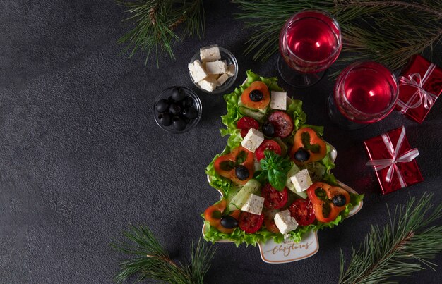 Insalata greca sana servita in un piatto come albero di Natale con decorazioni festive e due bicchieri di vino su sfondo scuro. Copia spazio