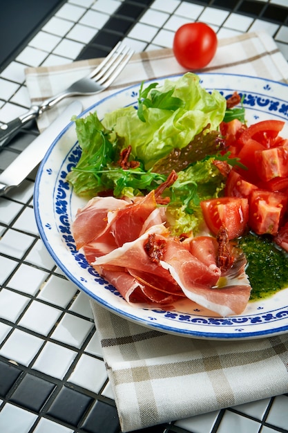 Insalata italiana deliziosa con il prosciutto di Parma, la salsa di pesto, la lattuga ed i pomodori ciliegia su un bello piatto ceramico sulla tavola bianca. Vista da vicino sul cibo