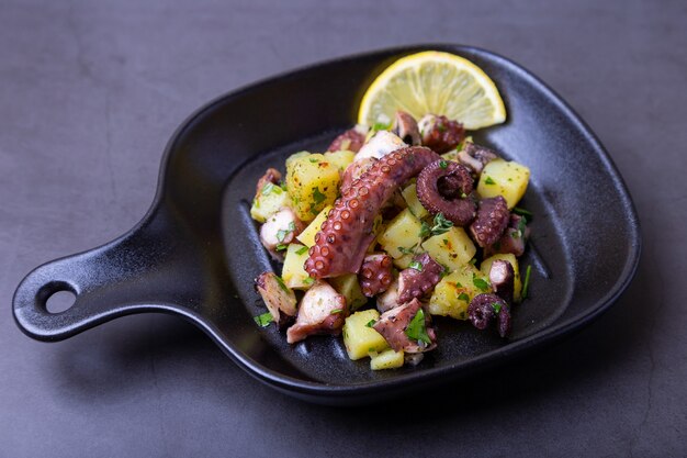 Insalata tiepida con polpo, patate e limone in un pentolino nero. Close-up, sfondo nero.