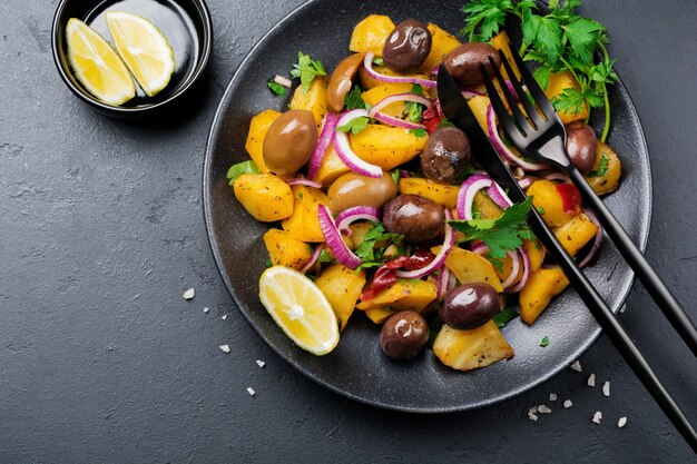 Insalata tiepida di patate con olive, pepe, prezzemolo e cipolla rossa su piatto in ceramica nera su superficie di cemento scuro