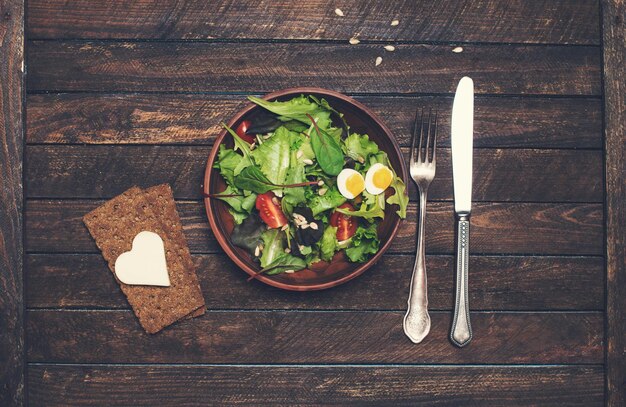 Insalata vegana fresca con patatine di pane formaggio a forma di cuore e argenteria su fondo di legno rustico