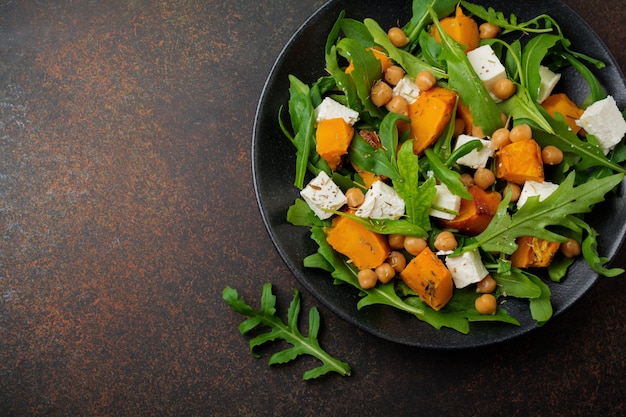 Insalata vegetariana con rucola, zucca al forno, erbe aromatiche e condimenti, formaggio feta, ceci e olio d'oliva in un piatto di ceramica nera su cemento scuro o pietra