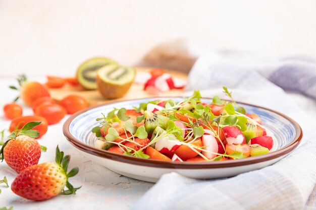 Insalata vegetariana di frutta e verdura di fragole, kiwi, pomodori, germogli microgreen su superficie di cemento bianco e tessuto di lino