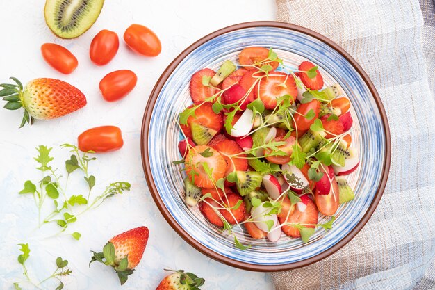 Insalata vegetariana di frutta e verdura di fragole, kiwi, pomodori, germogli microgreen su tavola di cemento bianco e tessuto di lino. Vista dall'alto, piatto laico, da vicino.