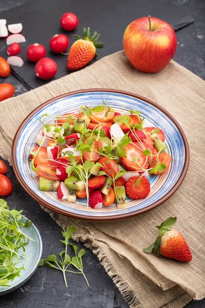 Insalata vegetariana di frutta e verdura di fragole, kiwi, pomodori, germogli microgreen su tavola di cemento nero e tessuto di lino. Vista laterale, da vicino.