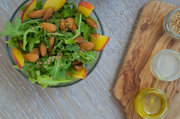 Insalata vitaminica - rucola con mandorle, pesche e semi di sesamo in un piatto di vetro. Olio d'oliva, succo di limone e semi di sesamo sul tagliere di legno sullo sfondo. Avvicinamento