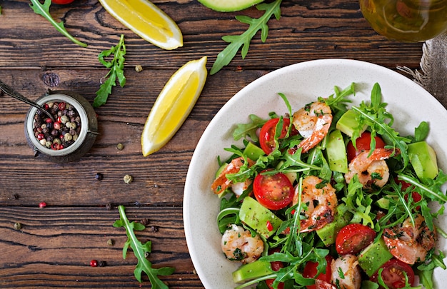 Insalatiera fresca con gamberetto, il pomodoro, l'avocado e la rucola sulla fine di legno della tavola su. Cibo salutare. Mangiare pulito. Vista dall'alto. Disteso.