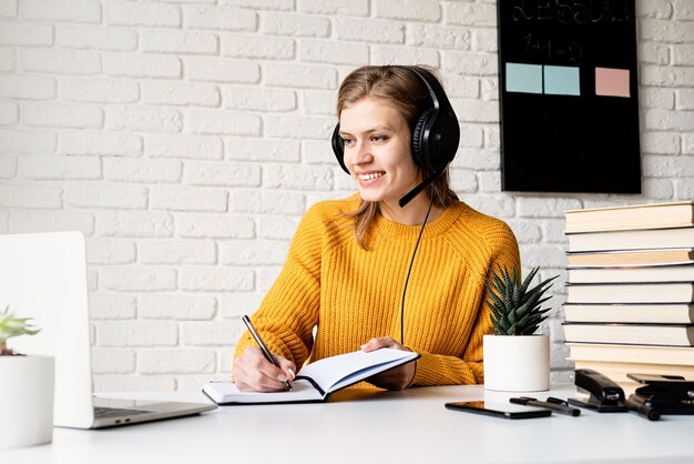 Insegnamento a distanza. E-learning. Giovane donna sorridente in maglione giallo e cuffie nere che studiano in linea facendo uso del computer portatile che scrive in taccuino