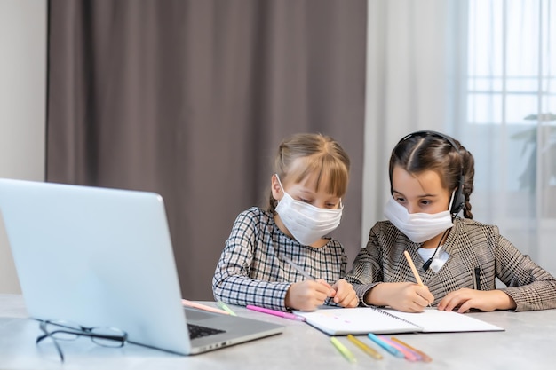 Insegnamento a distanza. Ragazze della scuola al computer portatile che indossano la maschera che studiano online Sedute al tavolo a casa