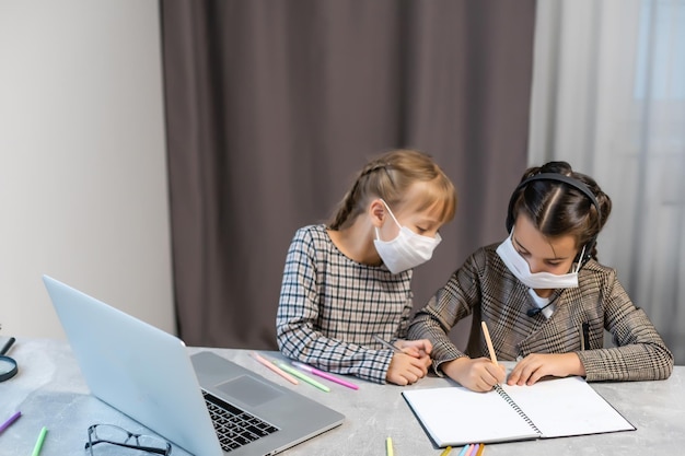 Insegnamento a distanza. Ragazze della scuola al computer portatile che indossano la maschera che studiano online Sedute al tavolo a casa