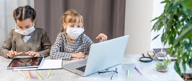 Insegnamento a distanza. Ragazze della scuola al computer portatile che indossano la maschera che studiano online Sedute al tavolo a casa