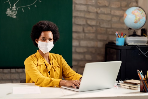 Insegnante afroamericano con una maschera facciale che lavora su un laptop in classe