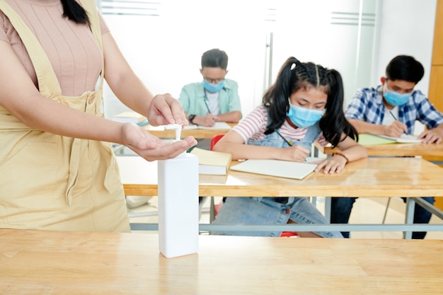 Insegnante che applica gel disinfettante sulle mani quando gli studenti in maschere mediche seduti alle scrivanie e scrivono in quaderni