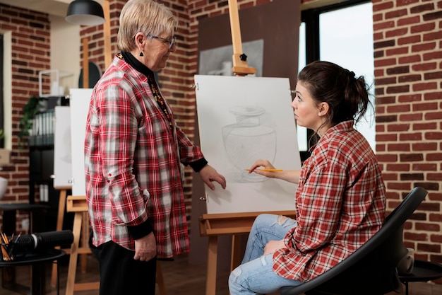 Insegnante di donna anziana sorridente amichevole in piedi vicino al cavalletto che lavora con lo studente durante la lezione di disegno. Figlia adulta che frequenta un corso d'arte di gruppo con la madre, trascorre del tempo insieme mentre impara a disegnare