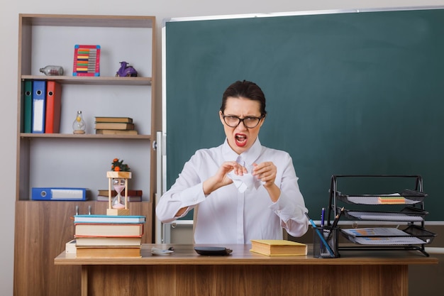 Insegnante di giovane donna che indossa occhiali strappando un pezzo di carta guardando scontento accigliato seduto al banco di scuola davanti alla lavagna in aula