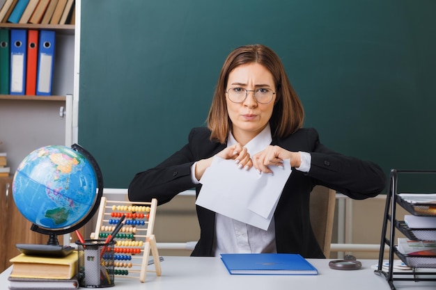 Insegnante di giovane donna con gli occhiali seduto al banco di scuola davanti alla lavagna in classe strappando un pezzo di carta con espressione aggressiva