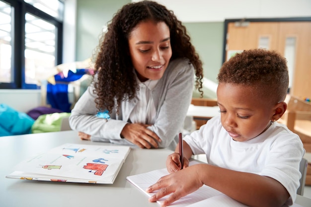 Insegnante di scuola materna femminile che lavora uno su uno con un giovane scolaro seduto a un tavolo che scrive in una vista frontale dell'aula da vicino