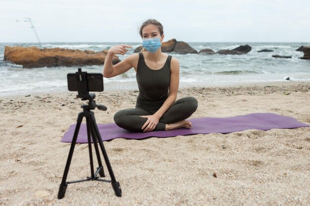 Insegnante di yoga di giovane donna in maschera medica conduce lezione online in spiaggia La donna sportiva trasmette alla fotocamera dello smartphone in piedi sul treppiede Blogging vlog Stile di vita sano