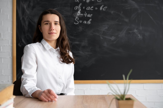 Insegnante donna millenaria seria in classe vicino alla lavagna della scuola con formule nella lezione Ragazza seduta a una scrivania al college Concetto di istruzione e studio Ritorno a scuola per il testo