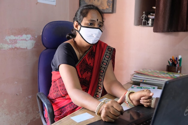 Insegnante femminile indiana in una maschera facciale che utilizza un computer portatile a scuola durante la pandemia di Covid-19