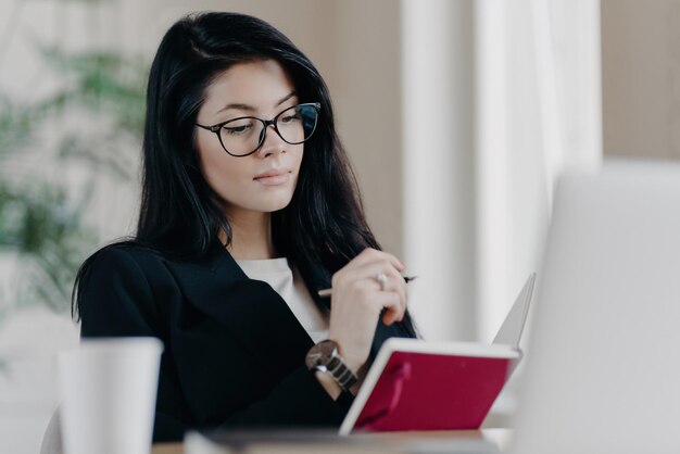 Insegnante femminile intelligente intelligente con gli occhiali per la correzione della vista apprende i corsi online prende appunti nel blocco note mentre il webinar degli orologi lavora in remoto pianifica le attività del progetto Concetto di occupazione delle persone