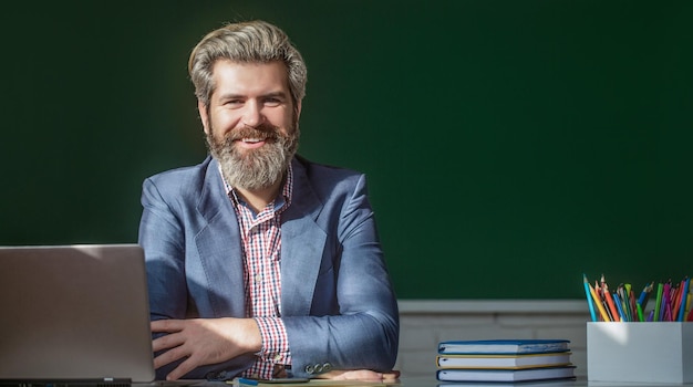 Insegnante sorridente in classe alla scuola elementare Ritratto di insegnante maschio fiducioso in classe Giovane insegnante maschio con laptop in classe si siede al tavolo la lavagna in classe