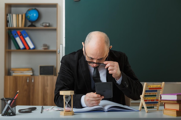 Insegnante uomo che indossa occhiali che controlla il registro di classe guardando da vicino la calcolatrice seduta al banco della scuola davanti alla lavagna in classe