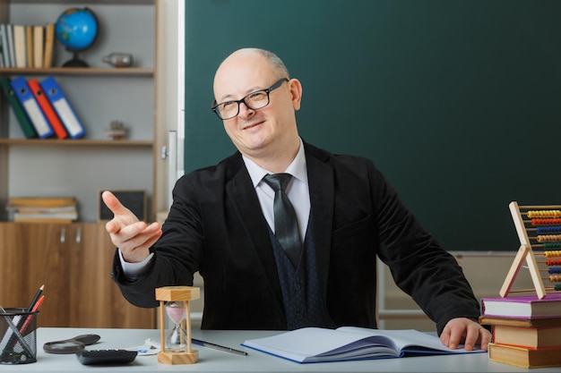 Insegnante uomo che indossa occhiali controllando il registro di classe guardando da parte felice e soddisfatto sorridente amichevole con il braccio alzato seduto al banco della scuola davanti alla lavagna in classe