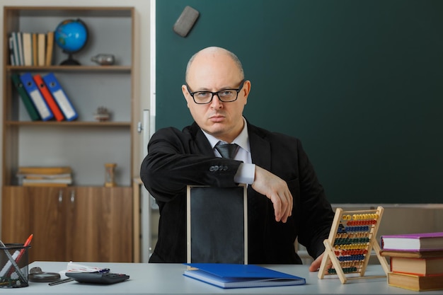 Insegnante uomo con gli occhiali seduto al banco di scuola davanti alla lavagna in classe che mostra lavagna che spiega la lezione che spiega la lezione con la faccia seria