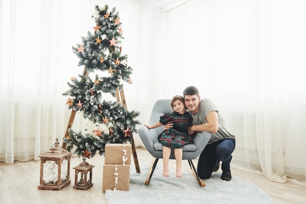 Insieme in sala. Concezione di Natale e festività. La bambina carina con suo padre è seduta sulla sedia vicino alla scala decorata con stelle e scatole regalo sul pavimento.