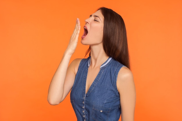 Insonnia, mancanza di sonno. Donna bruna stanca e annoiata in abito di jeans che sbadiglia ampiamente e copre la bocca con il braccio, sentendosi assonnata, pigra e affaticata. tiro in studio indoor isolato su sfondo arancione