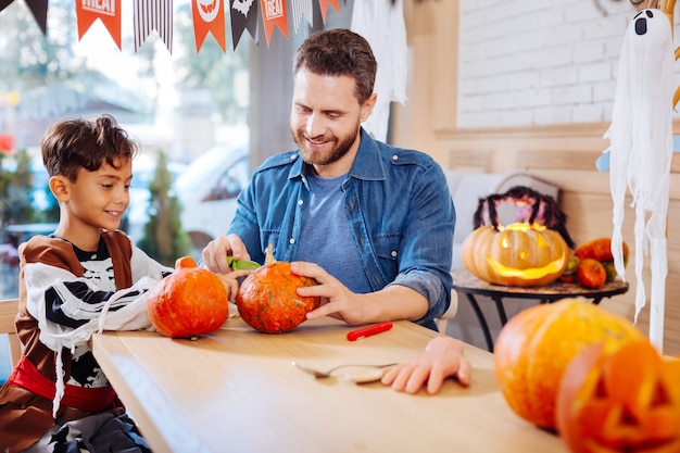 Intagliare la zucca. Simpatico figlio raggiante che indossa il costume di Halloween si sente allegro mentre intaglia la zucca con suo padre