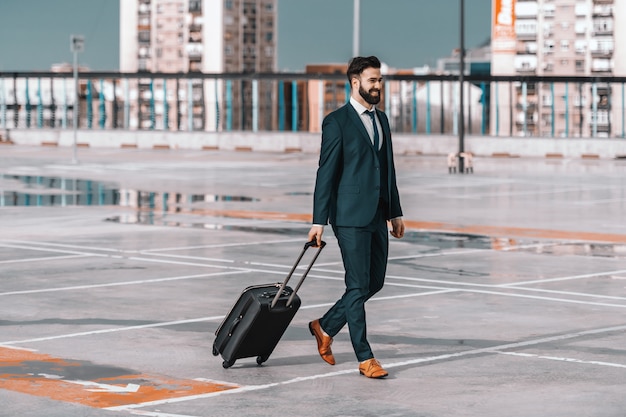 Integrale dell'uomo d'affari sorridente nell'usura convenzionale con bagagli che cammina sul parcheggio. Concetto di viaggio d'affari.