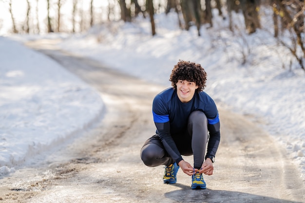 Integrale dell'uomo sportivo caucasico che si accovaccia all'aperto in abiti sportivi e che lega laccetto. Concetto di fitness invernale.