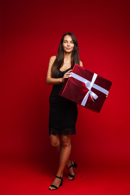 Integrale della giovane donna sorpresa in vestito nero che tiene grande contenitore di regalo, isolato sulla parete rossa