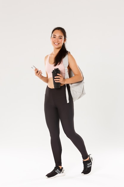 Integrale della ragazza asiatica sana e sottile sorridente che va allenamento di fitness, zaino per il trasporto atletico femminile con attrezzatura per l'allenamento e bottiglia d'acqua, utilizzando l'applicazione sportiva del telefono cellulare, sfondo bianco.