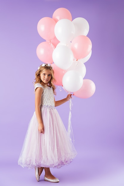 Integrale di una bella bambina vestita in abito da principessa in piedi isolato sopra la parete viola, tenendo in mano un mazzo di palloncini