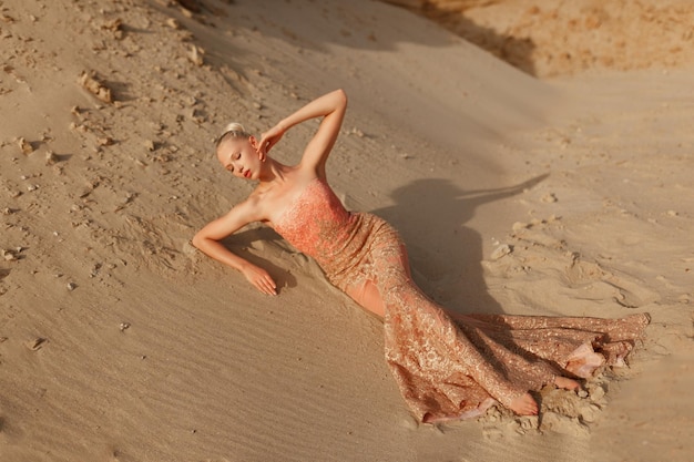 Integrale di una donna bionda in vestito d'oro costoso in posa sensuale nel deserto, sdraiata sulla sabbia al tramonto.