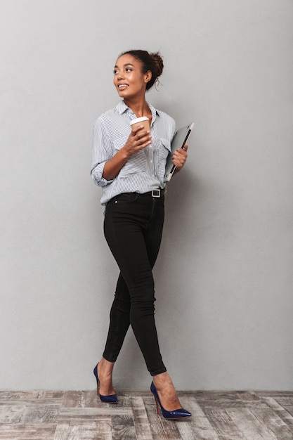 Integrale di una giovane donna africana attraente di affari che indossa la camicia che cammina isolata, tenendo il computer portatile, bevendo caffè da asporto