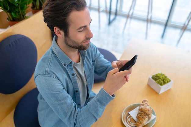 Interesse. Vista dall'alto del giovane uomo barbuto che guarda con interesse lo schermo dello smartphone seduto nella caffetteria al tavolo con uno spuntino