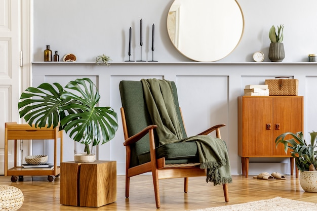 Interni eleganti del soggiorno con poltrona di design, comò vintage in legno, specchio rotondo, mensola, foglia tropicale, tavolino da caffè, decorazione, tappeto e accessori personali nell'arredamento della casa.