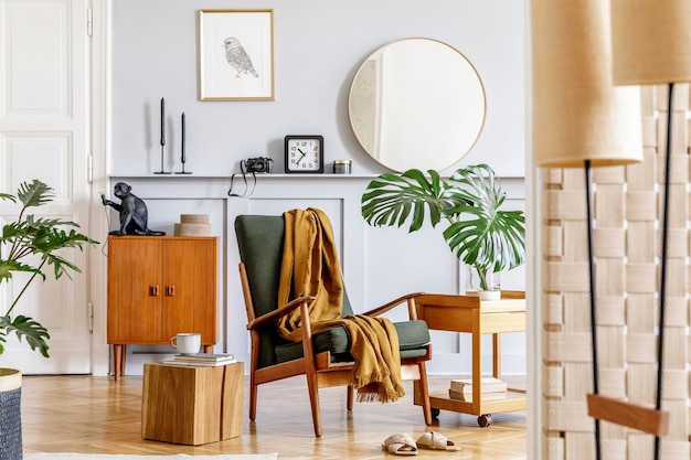 Interni eleganti del soggiorno con poltrona di design, comò vintage in legno, specchio rotondo, mensola, foglie tropicali, tavolino, decorazione, moquette e accessori personali nell'arredamento della casa.