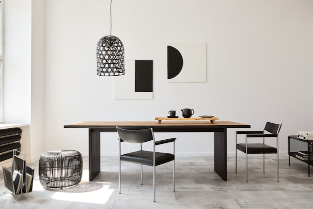 Interni eleganti della sala da pranzo con tavolo familiare in legno di design, sedie nere, teiera con tazza, finti quadri d'arte sul muro e accessori eleganti nell'arredamento moderno della casa. Modello.