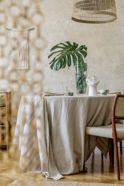 Interni eleganti della sala da pranzo con tavolo in legno, sedie di design, lampada a sospensione in rattan, fiori primaverili in vaso, bellissimi piatti, piante e decorazioni eleganti. Wabi sabi concetto..