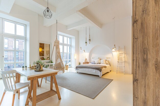 Interni moderni alla moda di un monolocale luminoso con colonne in legno in stile loft, decorato con mattoni, marmo e legno con mobili eleganti e pareti bianche