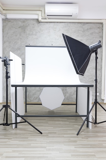 Interno con condizionatore d'aria studio bianco tavolo scena servizio fotografico e partizione con attrezzatura fotografica professionale softbox flash stroboscopico riflettore luce su treppiede pronto per i prodotti di ripresa.