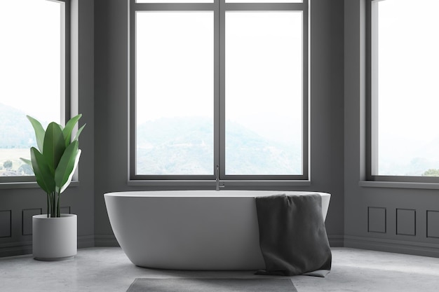 Interno di un bagno minimalista con pareti grigie, pavimento di cemento, grandi finestre e vasca da bagno bianca con asciugamano grigio.