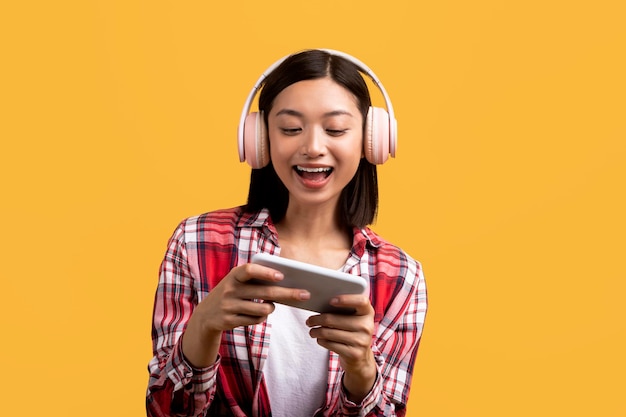 Intrattenimento e giochi online Gioiosa signora asiatica in moderne cuffie wireless che giocano su smartphone
