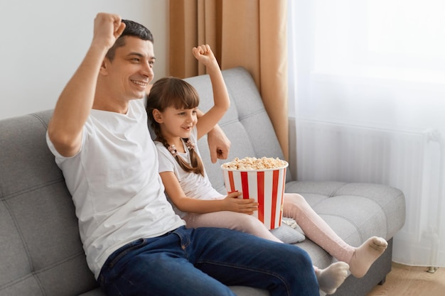 Intrattenimento per il fine settimana Padre felice e figlia piccola che guardano la tv o una ragazza di film seduta sul divano con cesto di popcorn vicino a papà sul divano che trascorrono il tempo libero insieme