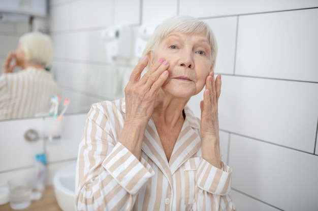 Invecchiamento. Donna dai capelli grigi guardarsi allo specchio in bagno e toccarsi il viso